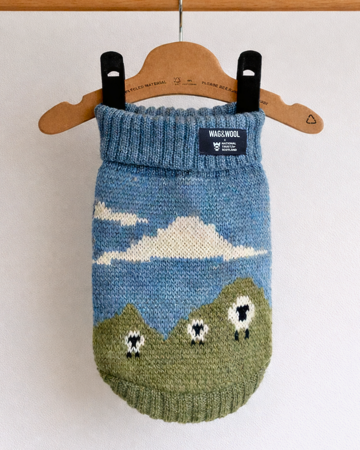 A knitted pet sweater in blue and green with a cloud and sheep motif, hanging from a wooden hanger on a wooden rod against a white wall.
