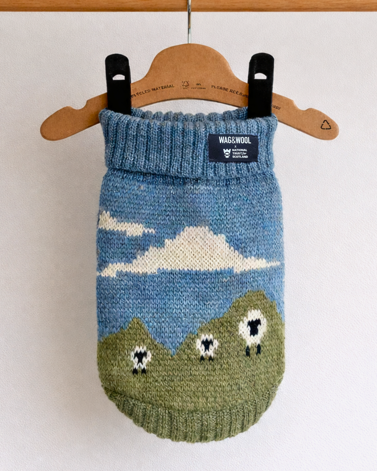 A knitted pet sweater in blue and green with a cloud and sheep motif, hanging from a wooden hanger on a wooden rod against a white wall.