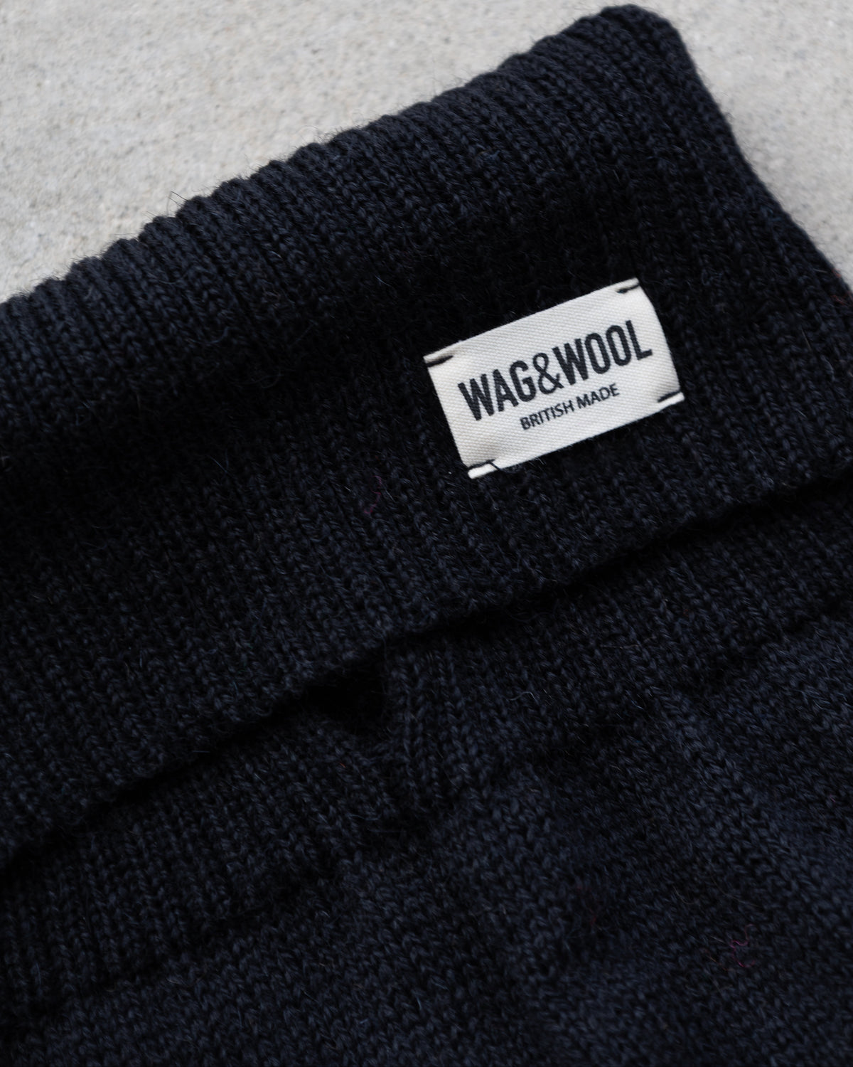 Close-up of a black knit dog jumper with a white sewn-in label reading WAG&WOOL BRITISH MADE, laid on a light gray textured surface.