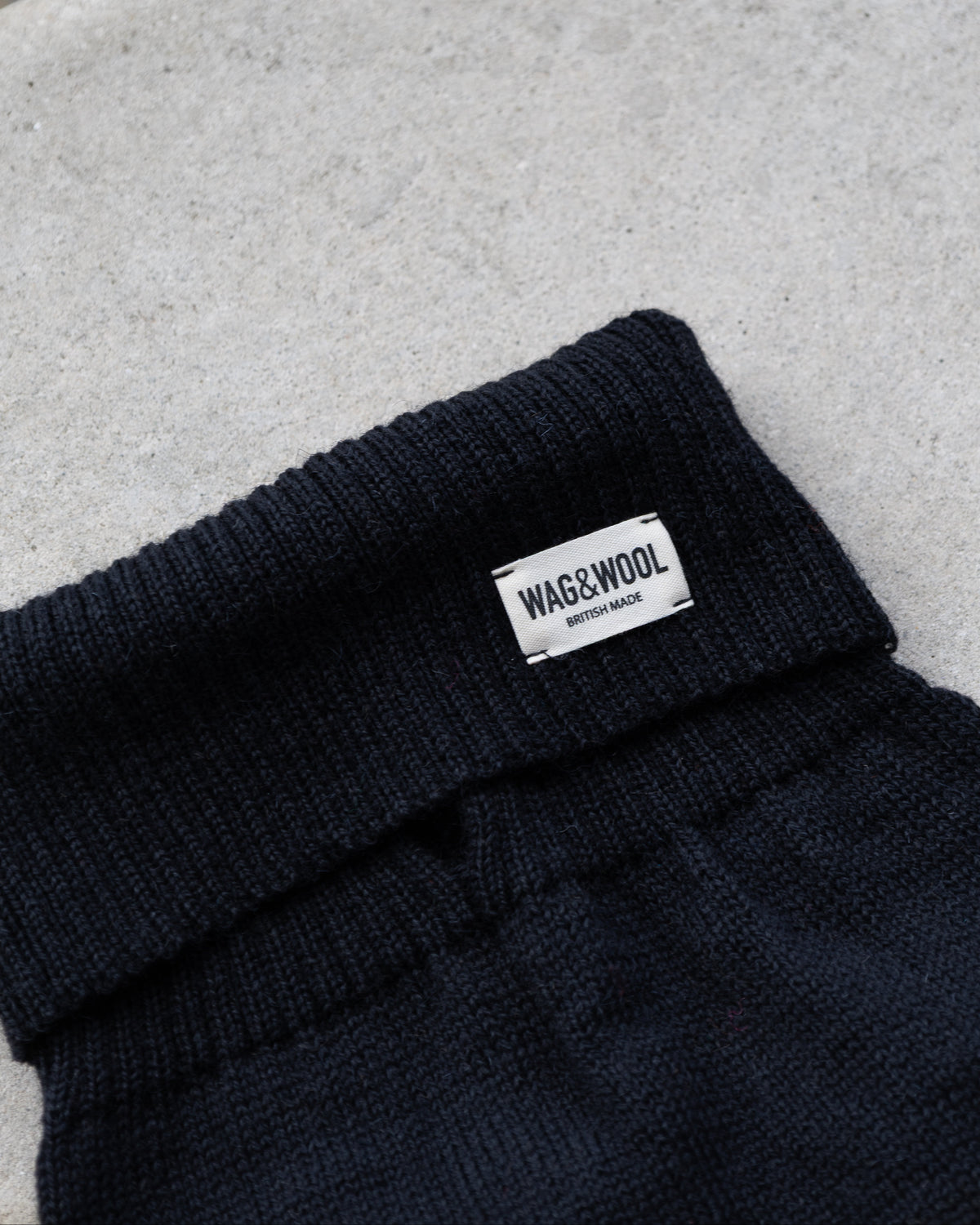 Close-up of a black dog jumper, with a small white rectangular label reading WAG&WOOL on the cuff, placed on a light concrete surface.