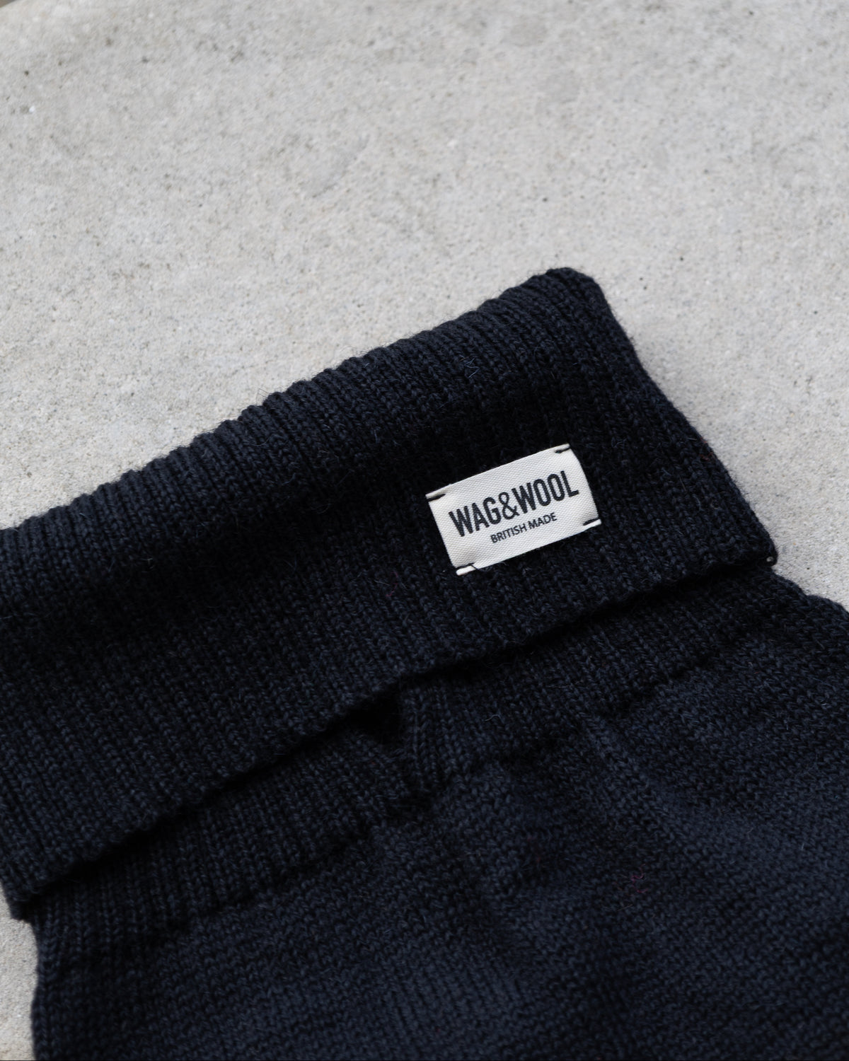 Close-up of a black dog jumper, with a small white rectangular label reading WAG&WOOL on the cuff, placed on a light concrete surface.