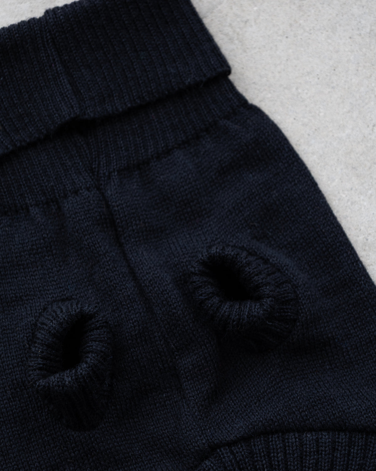 Close-up of a black knit pet sweater with two circular knit eye holes and a ribbed collar, laid flat on a light gray textured surface.
