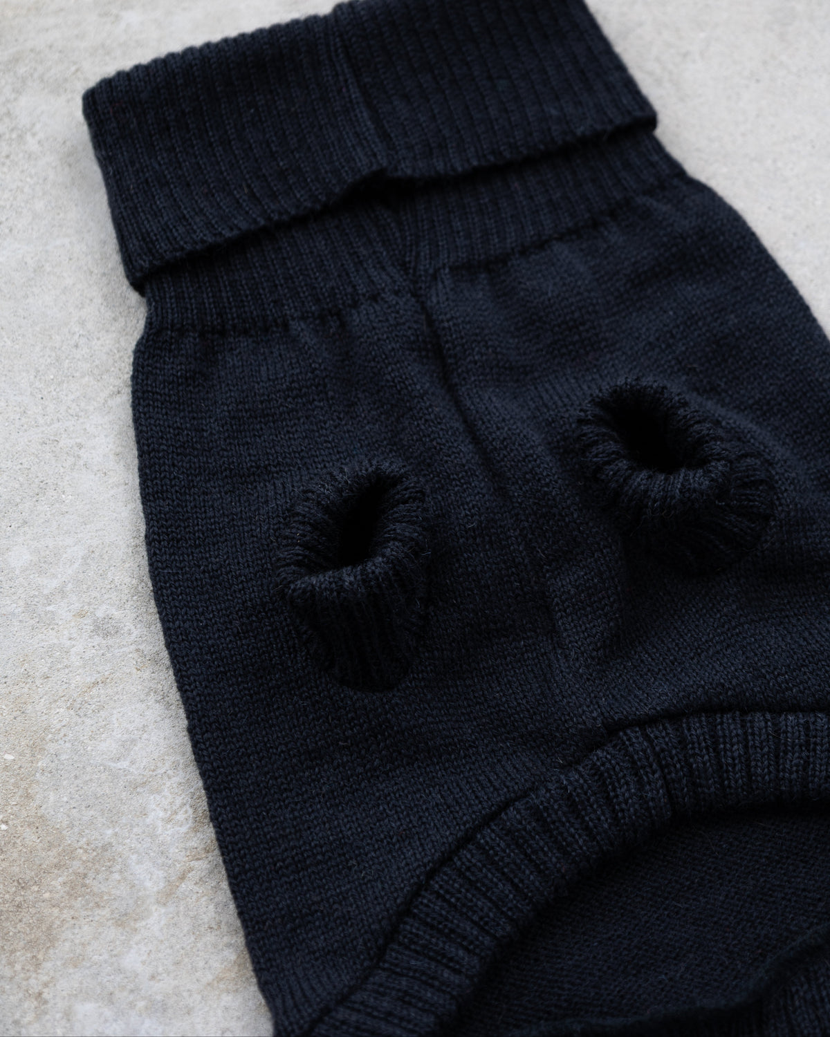 A close-up of a black knit pet sweater with two circular knit openings, laid on a light stone surface.