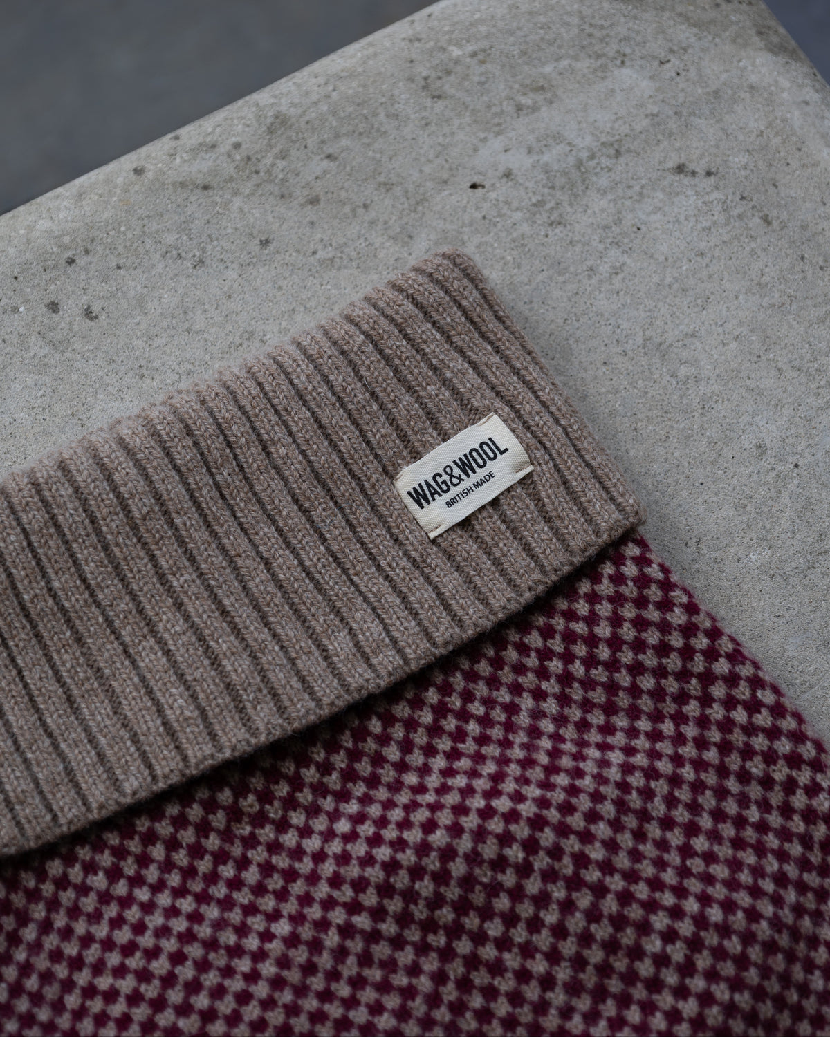 A close-up of a dog jumper with a small white tag reading wag and wool layered over a burgundy and cream check patterned fabric, set against a concrete surface.
