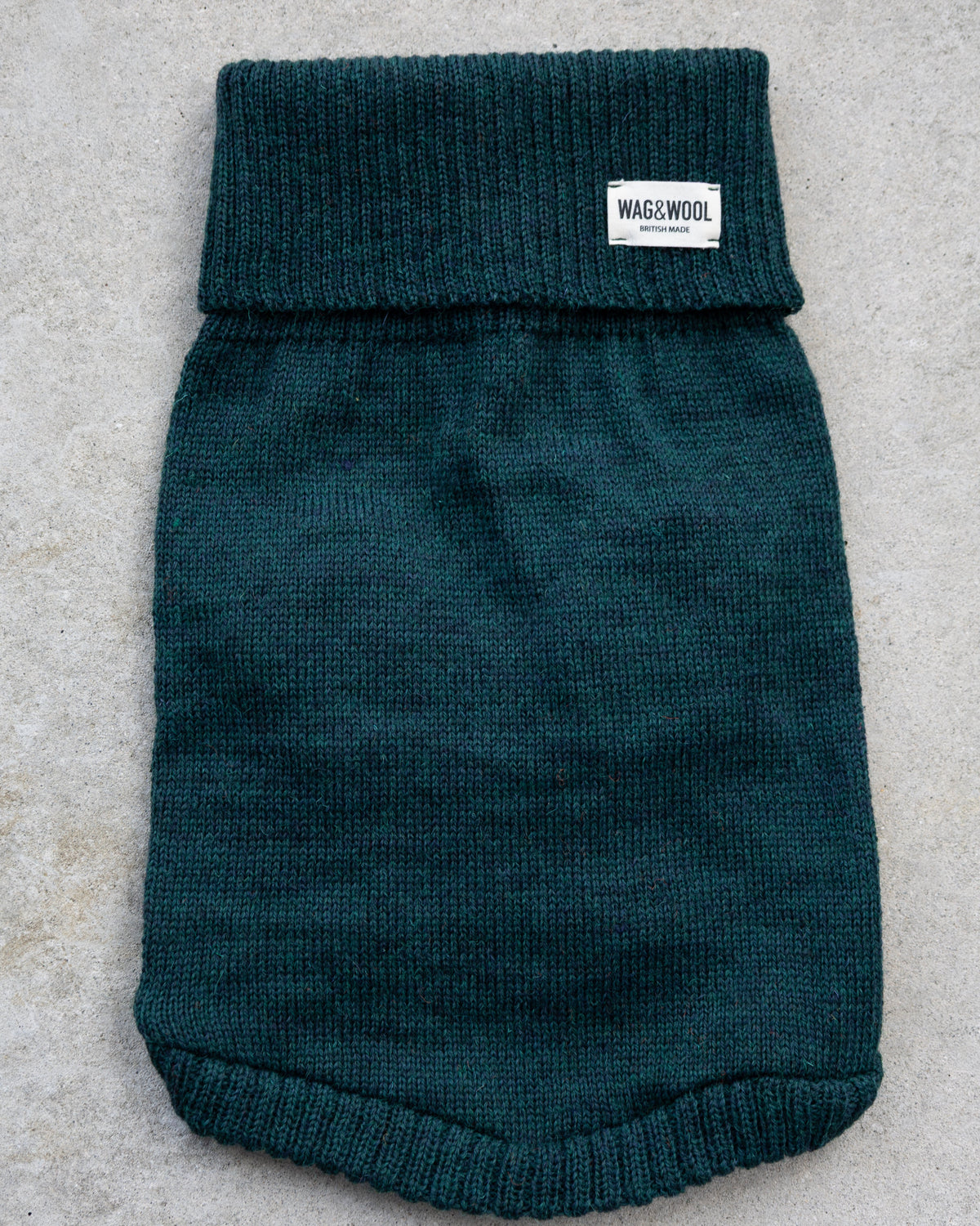 A dark green ribbed knit pet sweater or bootie with a white 'NAG&WOOL' brand label on the cuff, laid flat on a light gray textured surface.