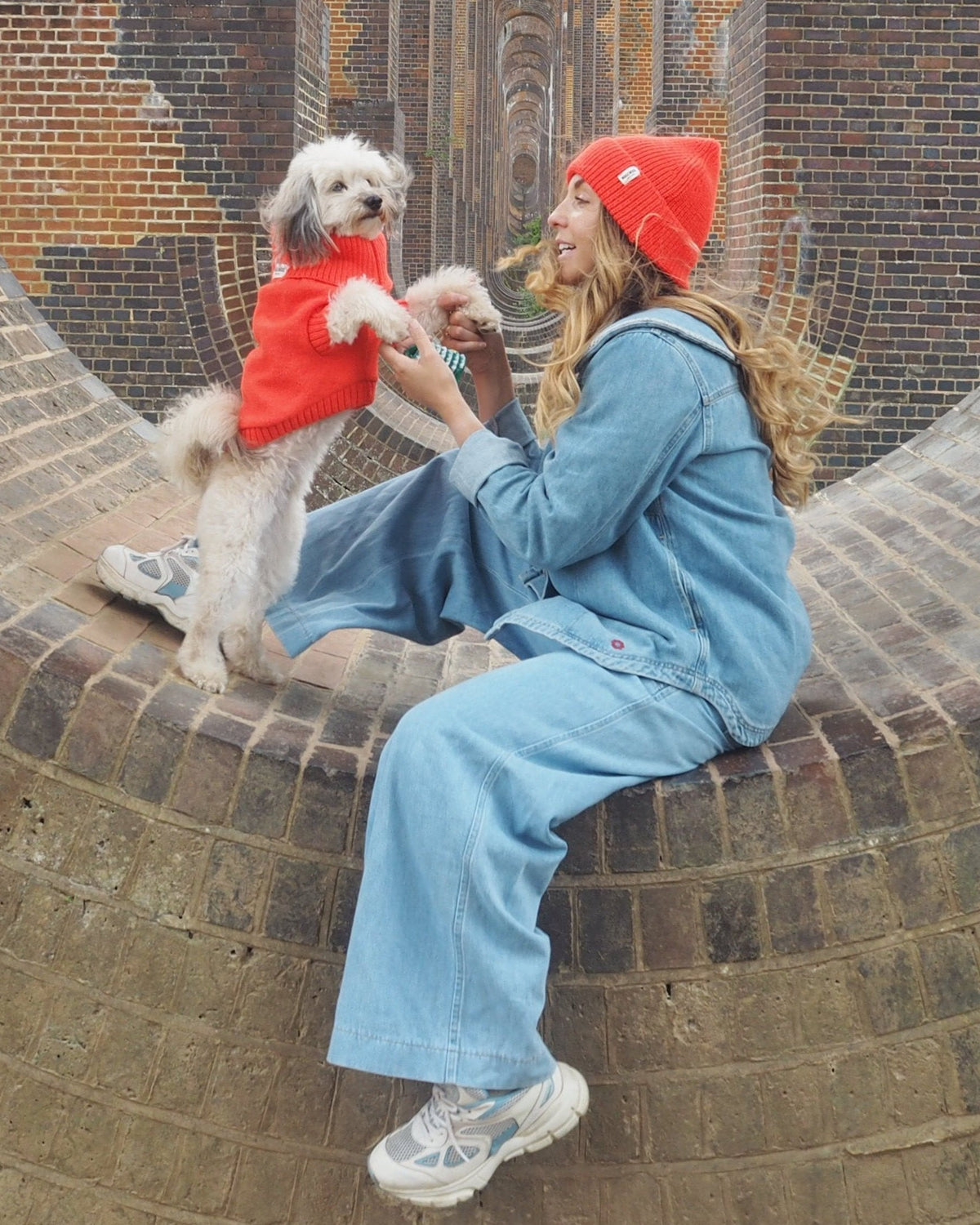 A person sitting on a brick structure with a small dog on their lap, both wearing matching lava orange jumpers.