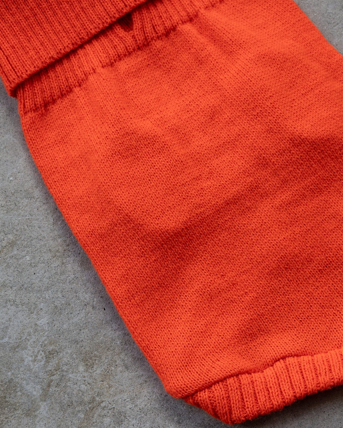 A close-up of a bright orange knit fabric, likely a dog sweater or garment, showing the ribbed cuff and hem against a textured gray concrete background.