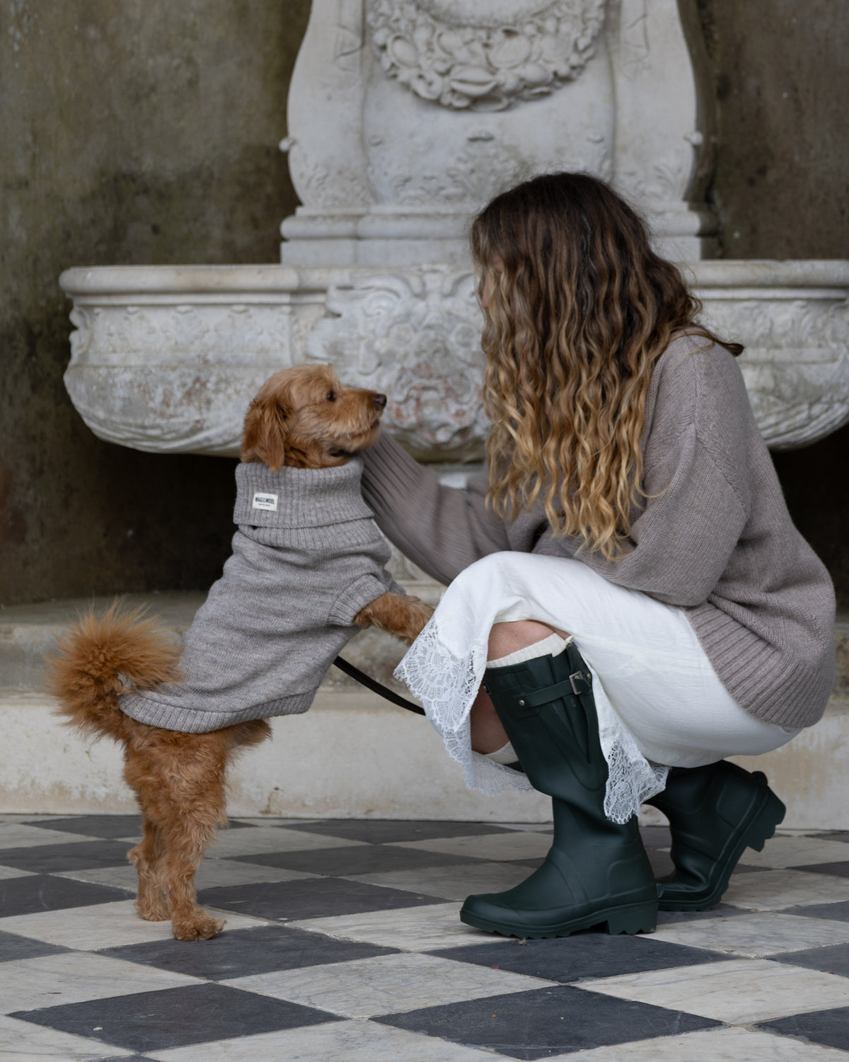 A woman crouches on a black-and-white checkered floor in front of an ornate stone fountain, wearing a taupe knit sweater, white skirt, and dark green rain boots. A small brown dog stands on its hind legs wearing a taupe knit sweater.