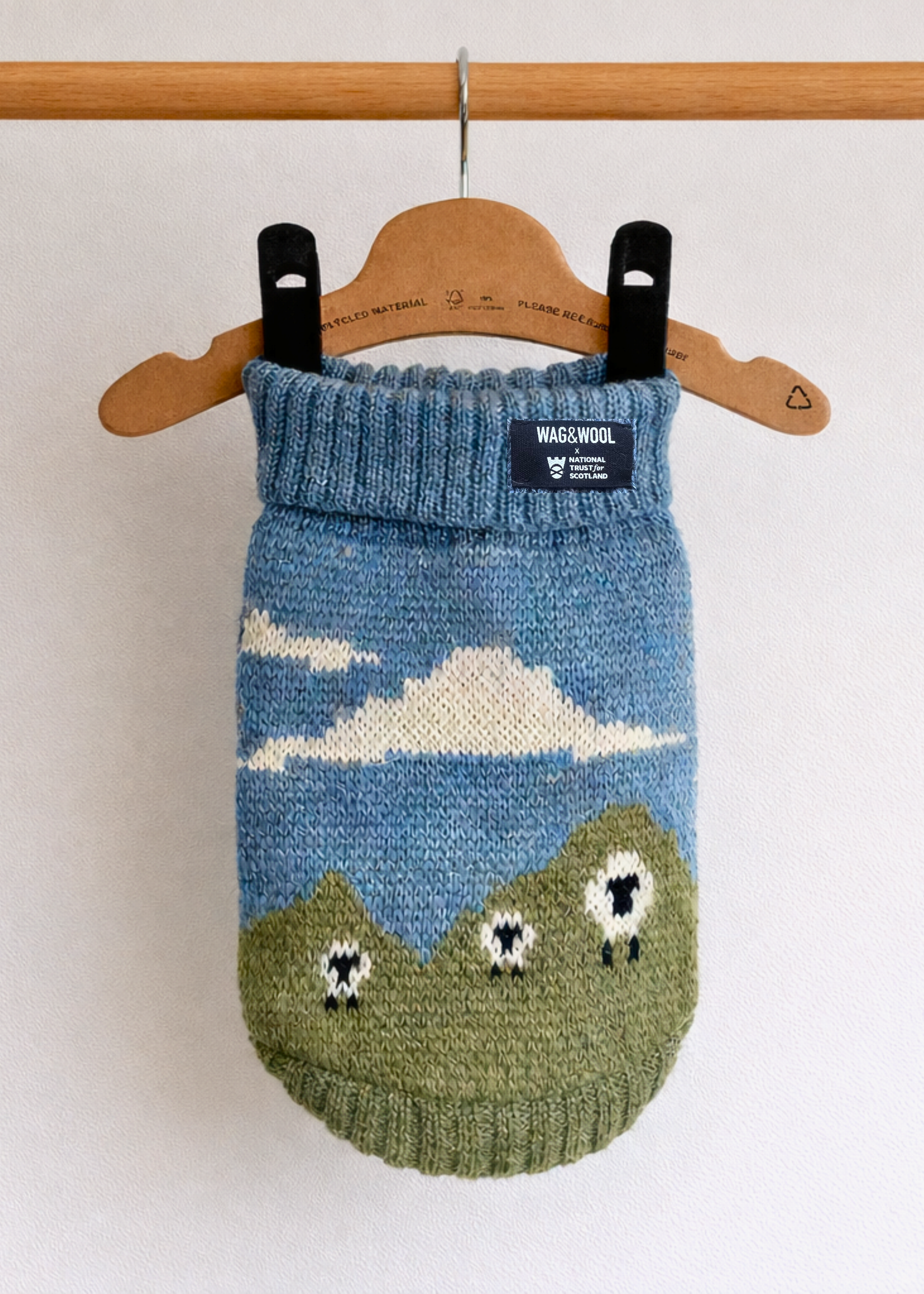 A knitted pet sweater in blue and green with a cloud and sheep motif, hanging from a wooden hanger on a wooden rod against a white wall. 