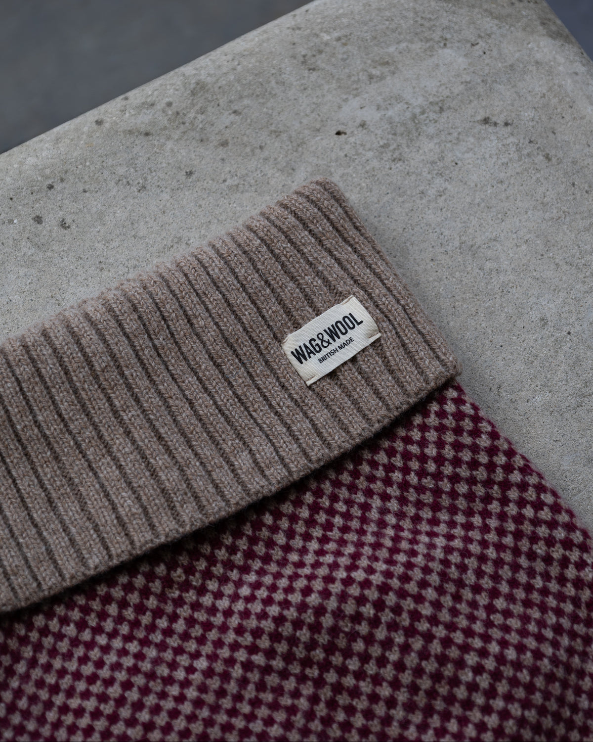 A close-up of a dog jumper with a small white tag reading wag and wool layered over a burgundy and cream check patterned fabric, set against a concrete surface.