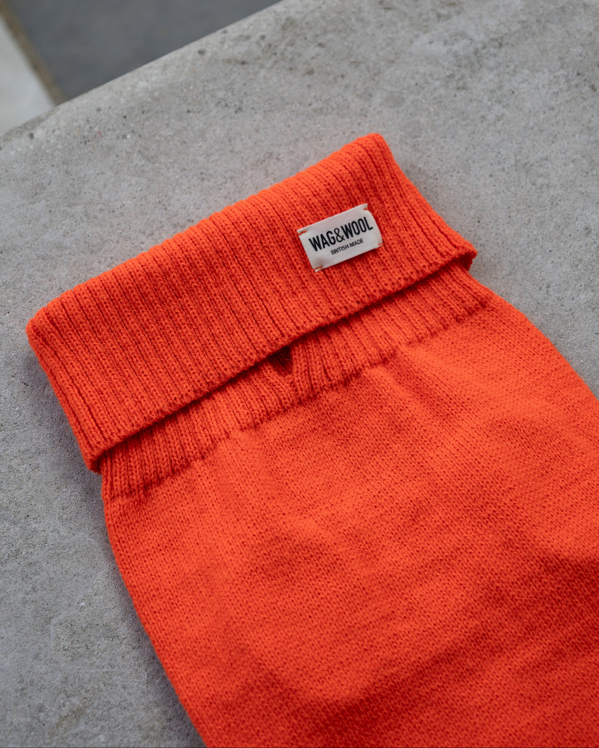 Bright orange knit dog sweater laid flat on a gray concrete surface, with a small white woven label reading WAGWOOL near the ribbed collar.