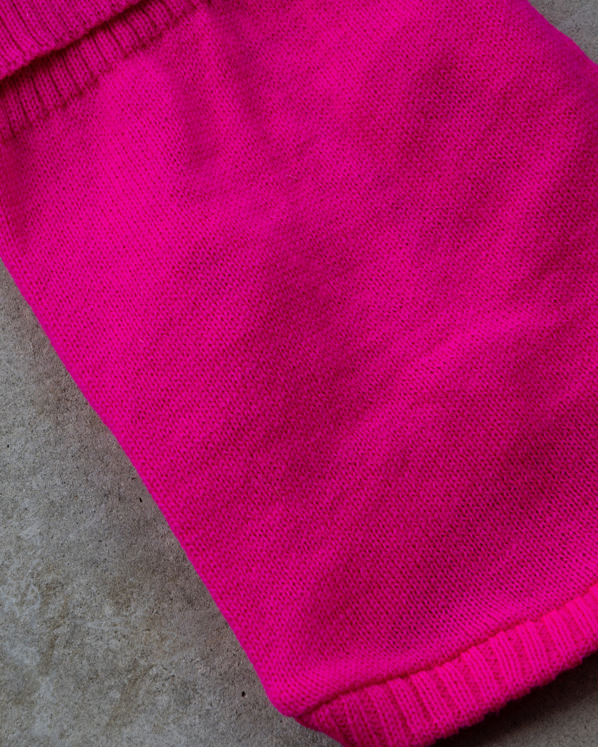 A close-up of a bright fuchsia pink dog jumper, showing the ribbed cuff and body texture, laid diagonally on a gray concrete surface.
